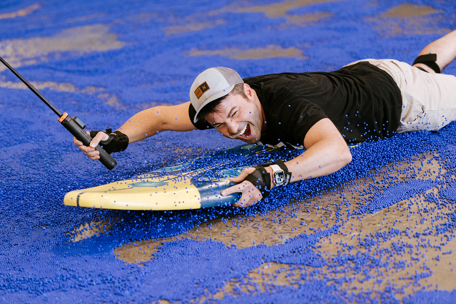 A man with fishing rod (Mark Rober) laying on surf board on top of a floor covered in blue metal pellets, giving a playful expression.