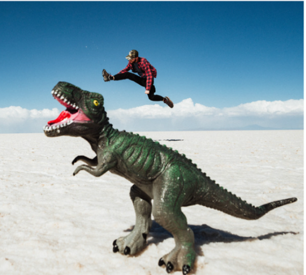 Person jumping over a toy dinosaur in a desert landscape. The perspective makes the person look smaller than the toy.