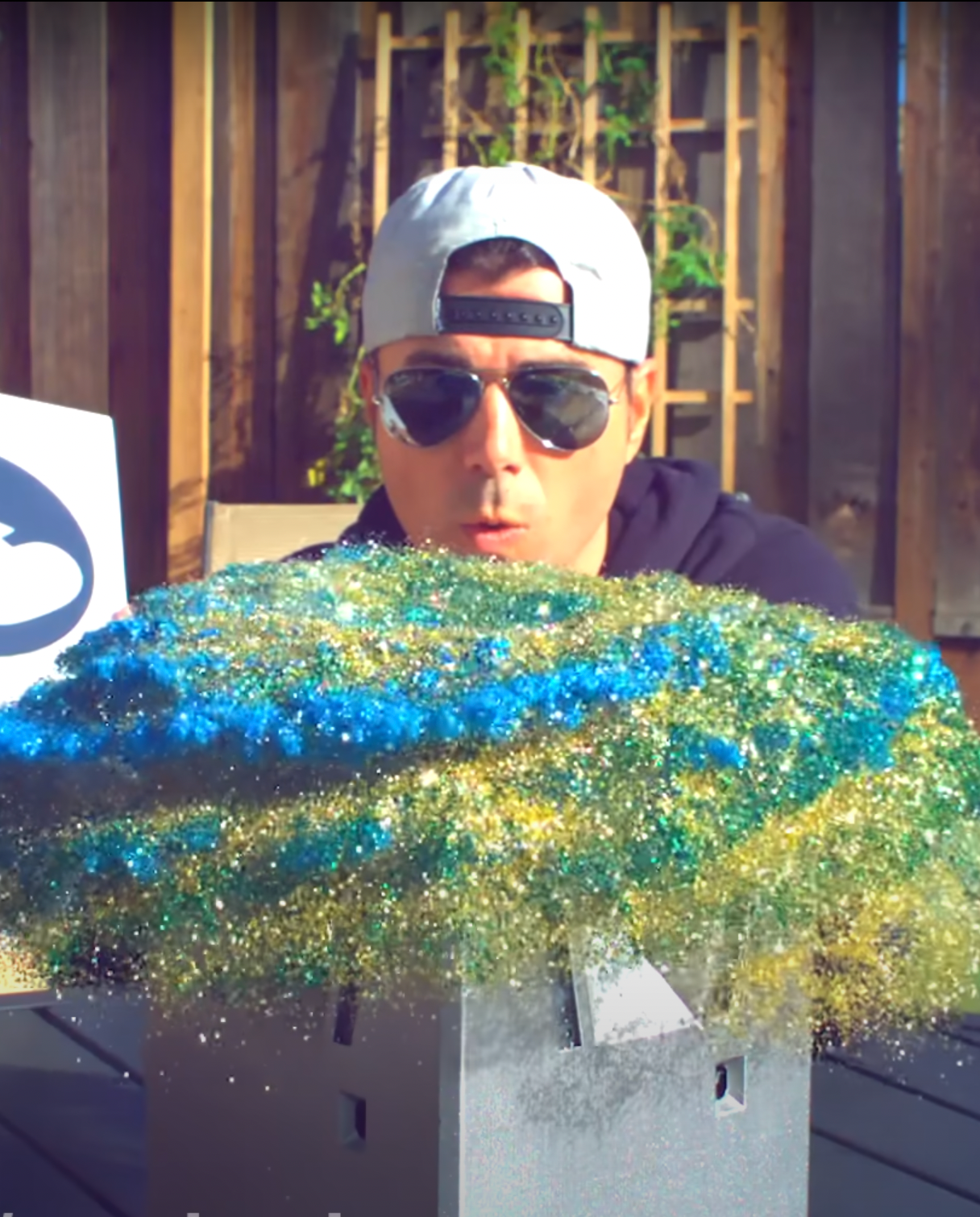 Mark Rober sits behind a glitterbomb. In front of him is a cloud of glitter.